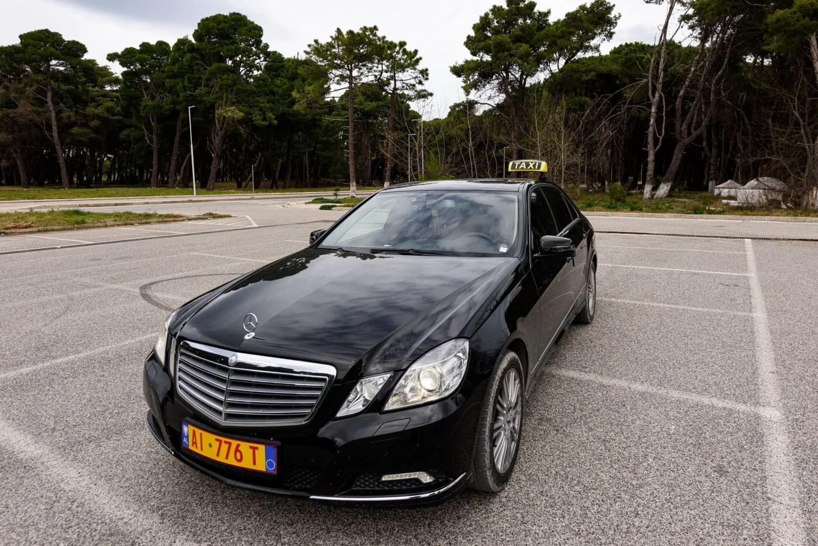 The black Mercedes-Benz E-Class of Dhermi Taxi, its yellow taxi roof sign lit, parked under the coastal pines near Dhërmi